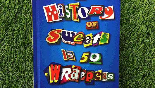 The cover of a blue book, with the title 'History of sweets in 50 wrappers' spelt out in colourful cut-out letters. The book is against a green background