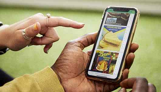 A close up of a hand holding a mobile phone.. On the mobile phone is a quiz with three pictures of desserts including cheesecake and madeira cake. There is another hand to the left with rings on pointing at the phone.
