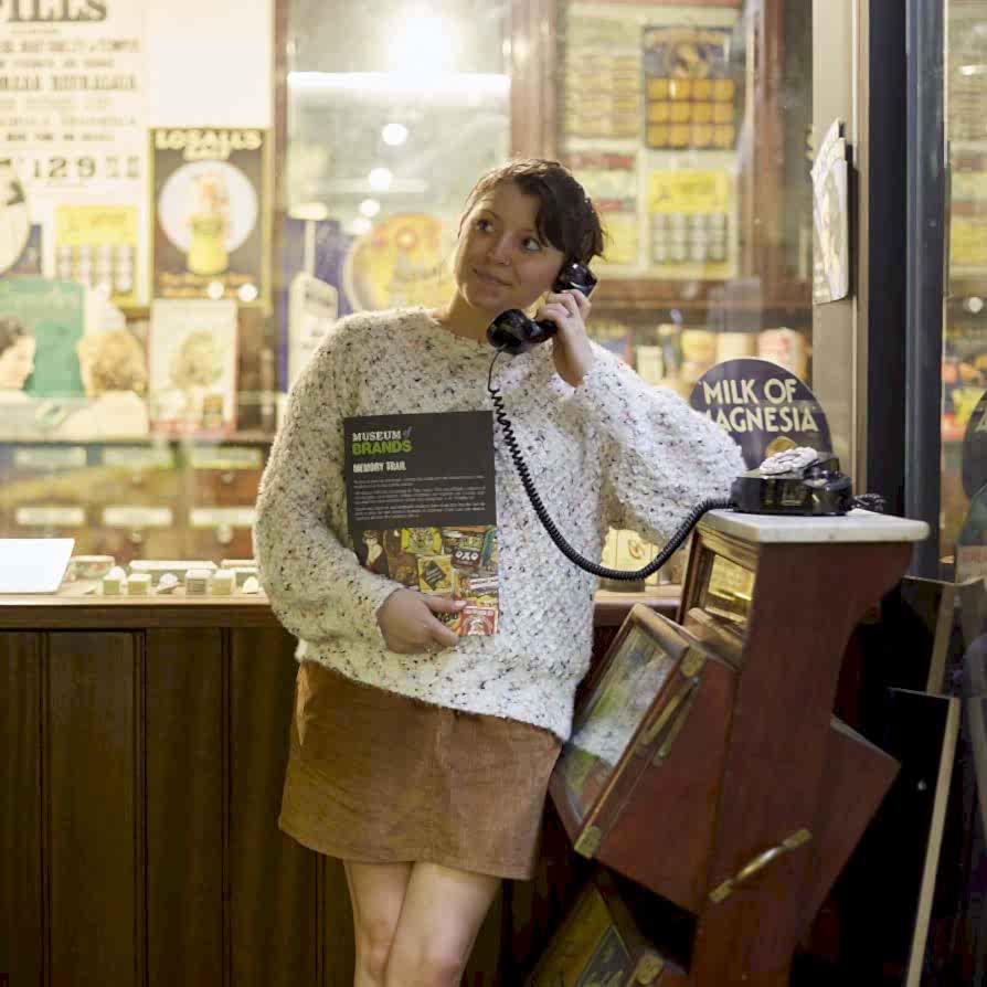 A woman in a white jumper and brown skirt stands holding a rotary telephone receiver to her ear. The telephone is an interactive part of the Museum collection that she is standing in