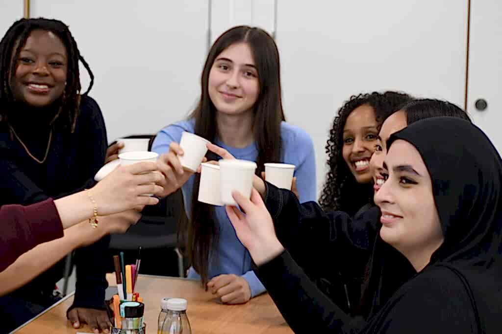 Five school students sit around a table smiling and toasting with cups of smoothie.