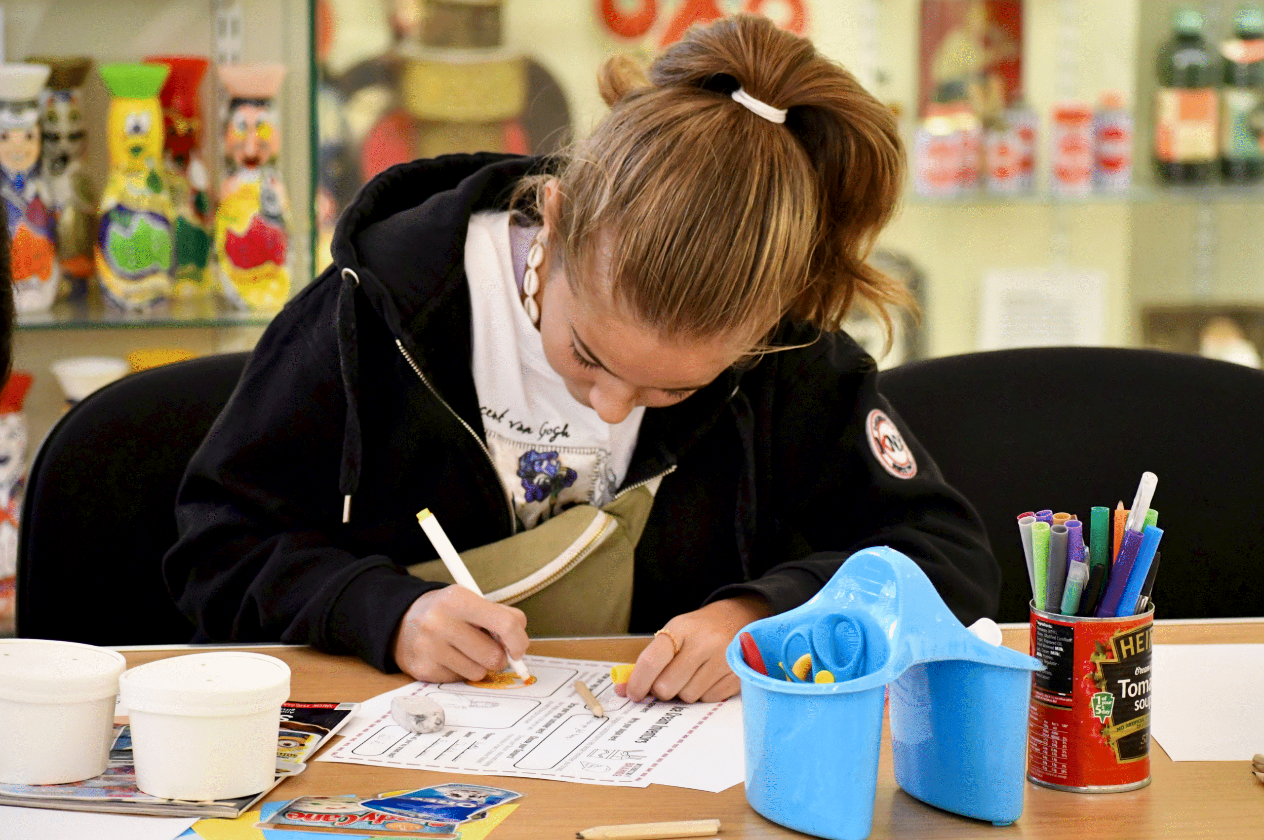 Young girl in a black hoodie sits at a table with craft supplies, writing on a worksheet